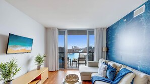 A modern living room with a sofa, a flat-screen TV, a dining table, and a large window with a city view.