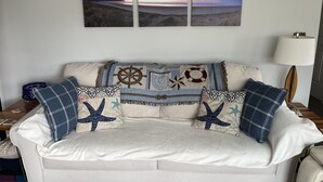 Uma sala de estar com um sofá decorado com almofadas de tema náutico, uma obra de arte pendurada na parede retratando uma cena de praia e um abajur sobre uma mesinha lateral.