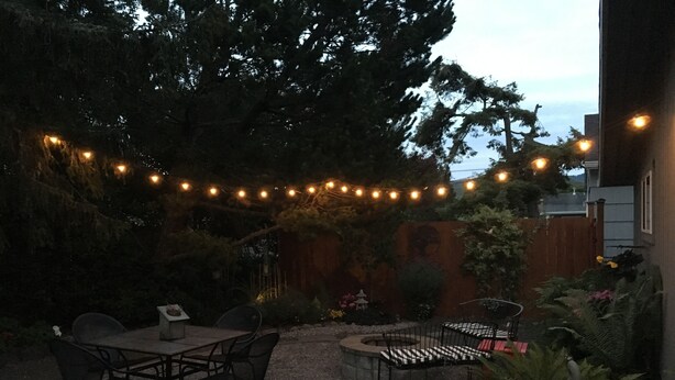 Uma área de pátio ao ar livre com mesa, cadeiras, luzes de festa e cerca de madeira.