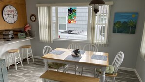 Uma área de refeições com uma mesa de madeira, cadeiras brancas e um banco. Há uma janela com persianas, um relógio e obras de arte nas paredes.