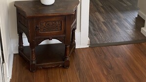 A wooden console table with a white textured vase and green plants, a decorative mirror above it, and a wooden floor leading to a kitchen area.