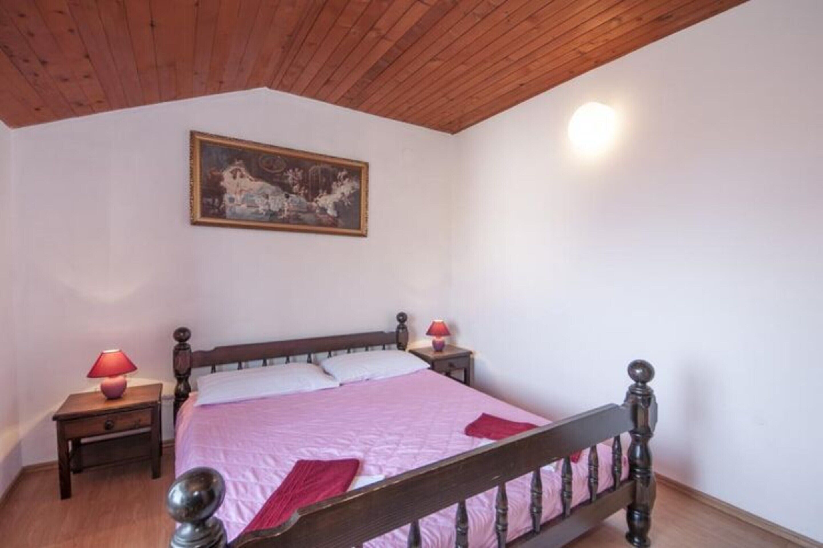 A bedroom with a wooden ceiling, a bed with a wooden headboard, two bedside tables with lamps, and a painting on the wall.