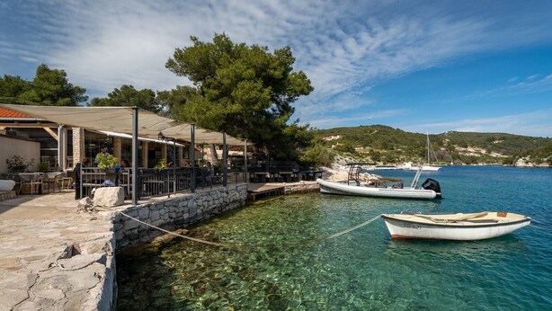 A coastal restaurant with outdoor seating, a stone wall, and boats in the water.