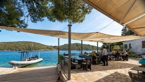 A coastal outdoor dining area with stone flooring, wooden tables, and a boat docked nearby.