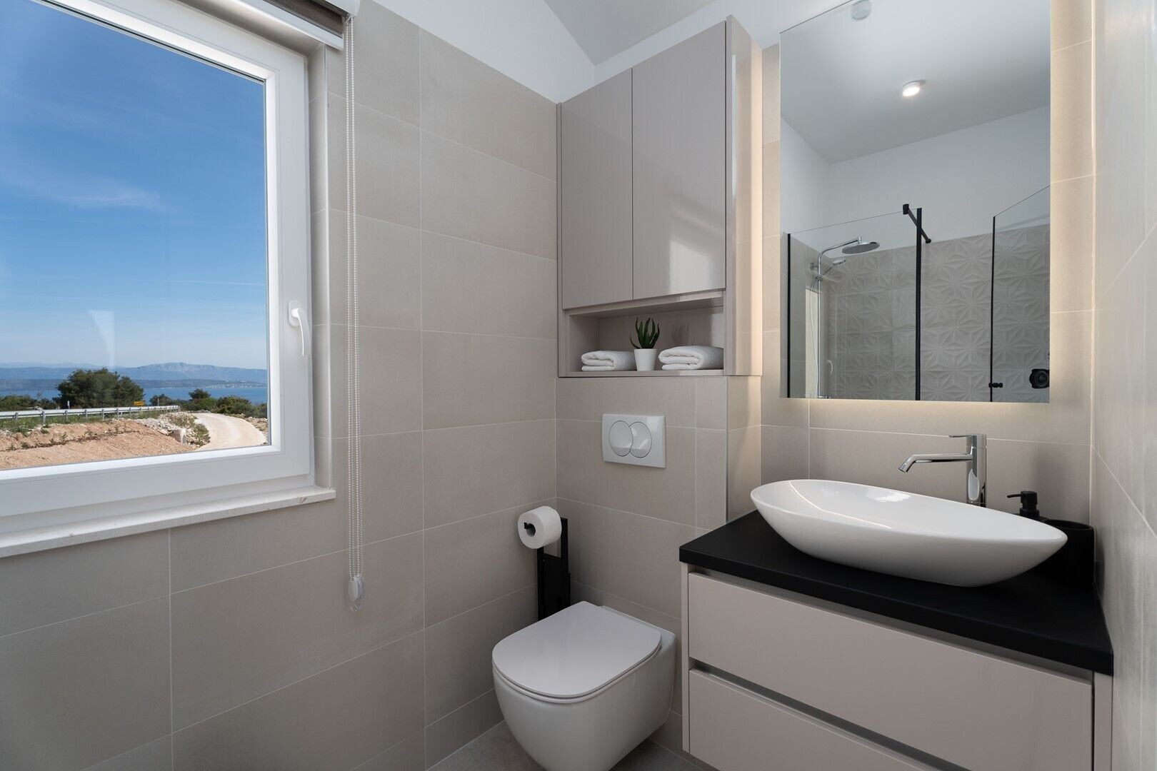 Un baño moderno con un ventanal grande, un inodoro blanco, una encimera negra con un lavabo blanco y un espejo sobre el lavabo.