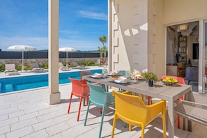 Uma área de refeições à beira da piscina, com uma mesa posta para dois, cadeiras coloridas e vista para a piscina e guarda-sóis ao fundo.