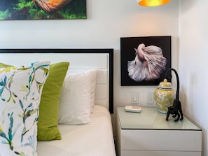En pænt redt seng med hvide og grønne puder, et indrammet kunstværk over sengen, et natbord med en lampe og en figur, samt en stikkontakt på væggen.