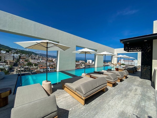 A rooftop pool area with lounge chairs and umbrellas, overlooking a cityscape.