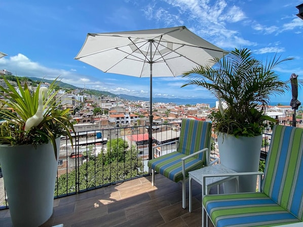 A balcony with a view of a cityscape, a striped chair, a potted plant, and a large umbrella. - Blissful Ocean-View Penthouse with Rooftop Pool (Puerto Vallarta)