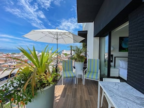 A balcony with a white umbrella, two striped chairs, a potted plant, and a view of a cityscape.