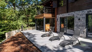 A modern outdoor patio with lounge chairs, a wooden deck, and a stone wall. - Chalet in the Woods With Nordic Bath (Manhay)