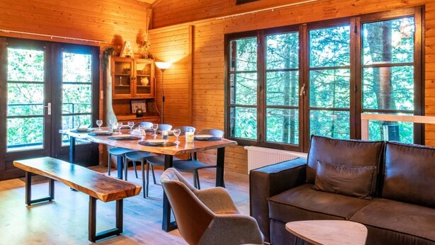 A cozy cabin interior with a dining area, a sofa, and large windows offering a forest view. - Chalet in the Woods With Nordic Bath (Manhay)
