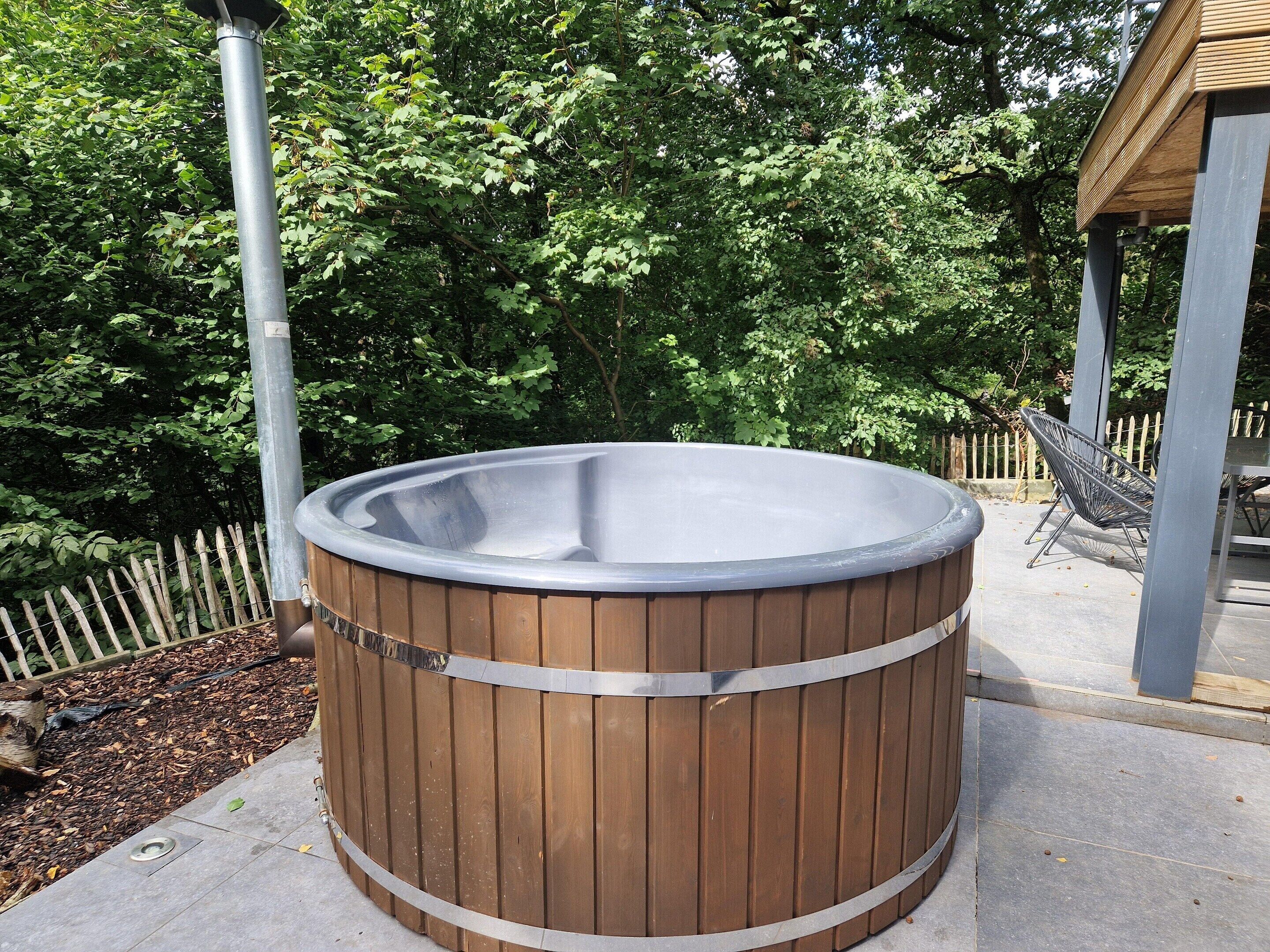 Een hottub met een houten buitenkant, omgeven door een houten omheining en bomen.