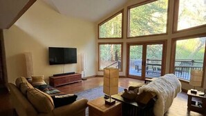 A spacious living room with a high ceiling, large windows, a ceiling fan, a flat-screen TV, and a cozy fireplace area. - Redwood Creek Getaway! (Redway)