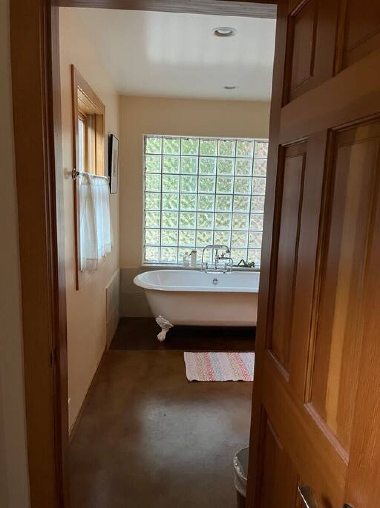 A narrow hallway leading to a bathroom with a freestanding bathtub, a window with a grid pattern, and a wooden door.