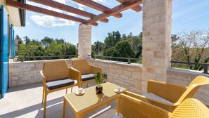 Un espace extérieur abrité, aménagé avec des meubles en osier, une table sur laquelle se trouvent une tasse et une soucoupe, et offrant une vue sur les arbres et le ciel.