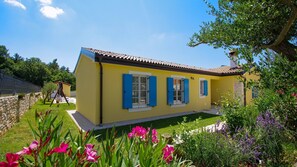 Une maison jaune aux volets bleus, entourée d’un jardin où poussent des fleurs et des arbres.