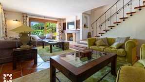 Ein geräumiges Wohnzimmer mit einer Couch, einem Sessel und einem Glastisch. Im Hintergrund ist eine Treppe mit Holzstufen und einem schwarzen Geländer zu sehen.