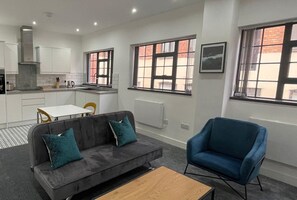 A compact living space with a kitchenette, a grey sofa with teal cushions, a blue armchair, and a wooden coffee table.