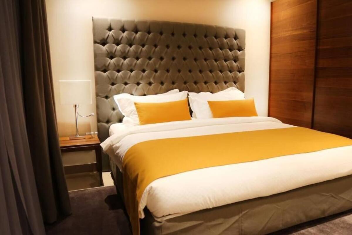 A hotel room with a tufted headboard, a bed with white and mustard bedding, and a wooden wardrobe.