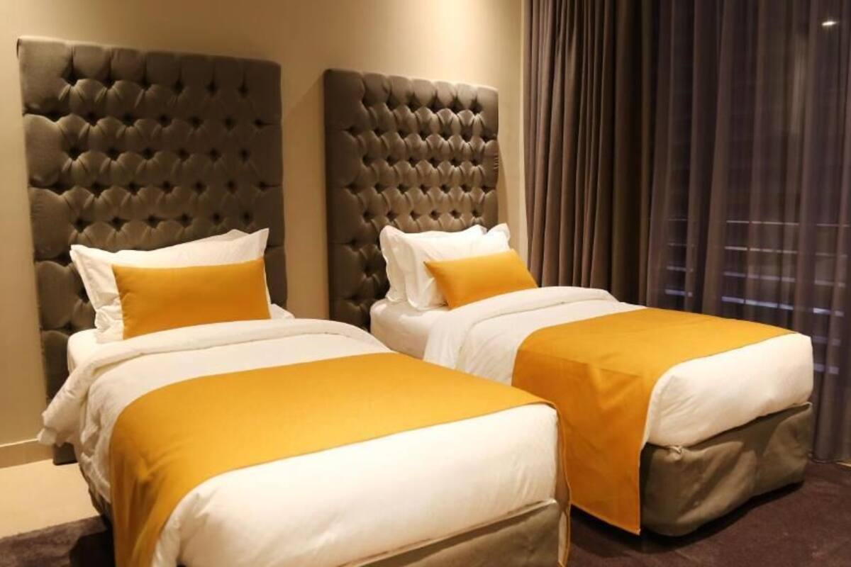 A hotel room with two beds, each with a tufted headboard, white bedding, and a mustard-colored throw.