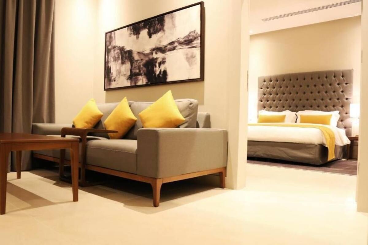 A modern hotel room with a grey sofa, wooden coffee table, and a large bed with a tufted headboard.