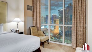 Habitación de hotel con una cama grande, una silla, una lámpara y vistas de la ciudad a través de amplias ventanas.