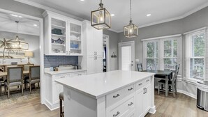 A modern kitchen with white cabinetry, a central island, pendant lighting, and a dining area with chairs and a table. - Award-Winning Resort Amenities | Grand Pavilion 71 by Wild Dunes (Isle of Palms)
