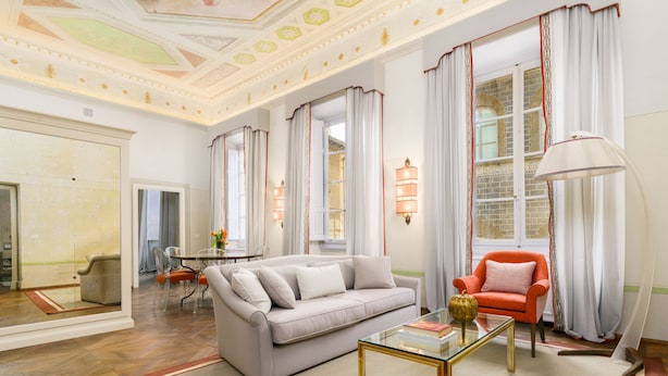 Un salon spacieux comprenant un canapé, un fauteuil et une table basse. On y trouve également un coin repas avec une table et des chaises. La pièce est dotée de grandes fenêtres avec des rideaux et d’un lustre.