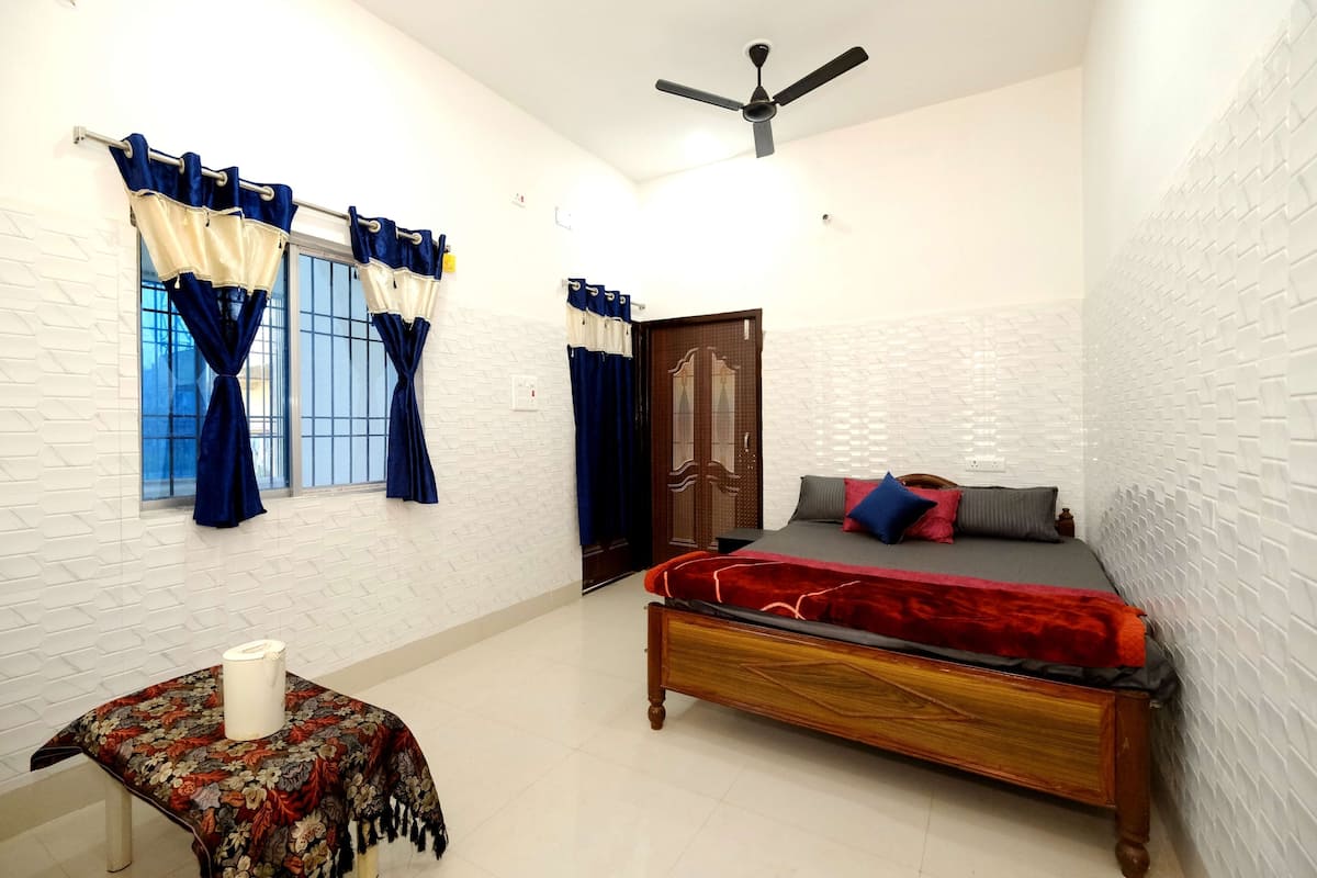 A bedroom with a bed, a table with a tablecloth, a ceiling fan, and windows with curtains.