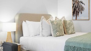 Ein ordentlich bezogenes Bett mit weißer Bettwäsche und gemusterten Kissen, eine Nachttischlampe und ein gerahmtes Bild von einer Palme an der Wand.