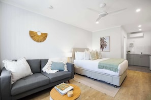 Un dormitorio moderno con un sofá gris, una cama con una colcha verde, un ventilador de techo y una zona de cocina con armarios blancos.