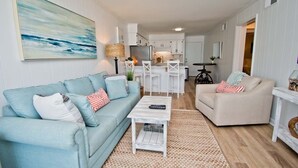 Ein Wohnzimmer mit einem hellblauen Sofa, einem beigen Sessel und einem Couchtisch. Im Hintergrund befindet sich eine Küche mit weißen Schränken und ein Essbereich mit weißen Stühlen.