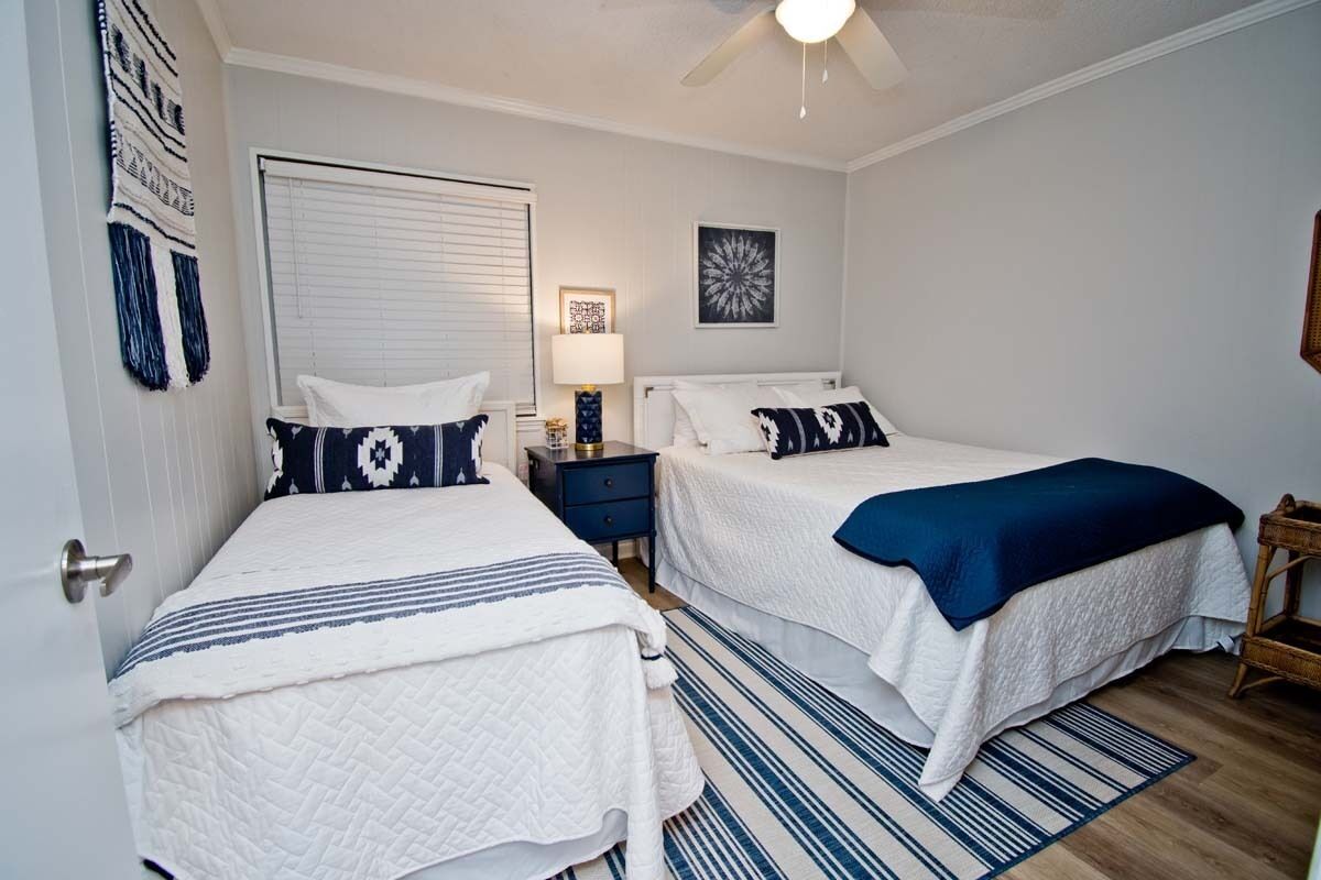 A bedroom with two beds, a ceiling fan, a window with blinds, a bedside table with a lamp, and a decorative wall hanging.