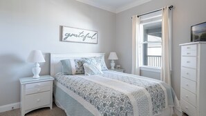 Ein Schlafzimmer mit einem Bett, zwei Nachttischen, einer Kommode, einem Fenster mit Vorhängen und einem an der Wand befestigten Schild.