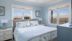 Un dormitorio con una cama, dos mesitas de noche, un ventilador de techo y amplios ventanales con vistas al paisaje.