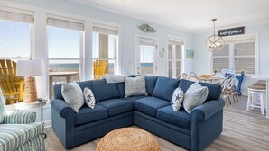 Una sala de estar con un sofá seccional azul, un otomano redondo tejido, y un comedor con una mesa y sillas.
