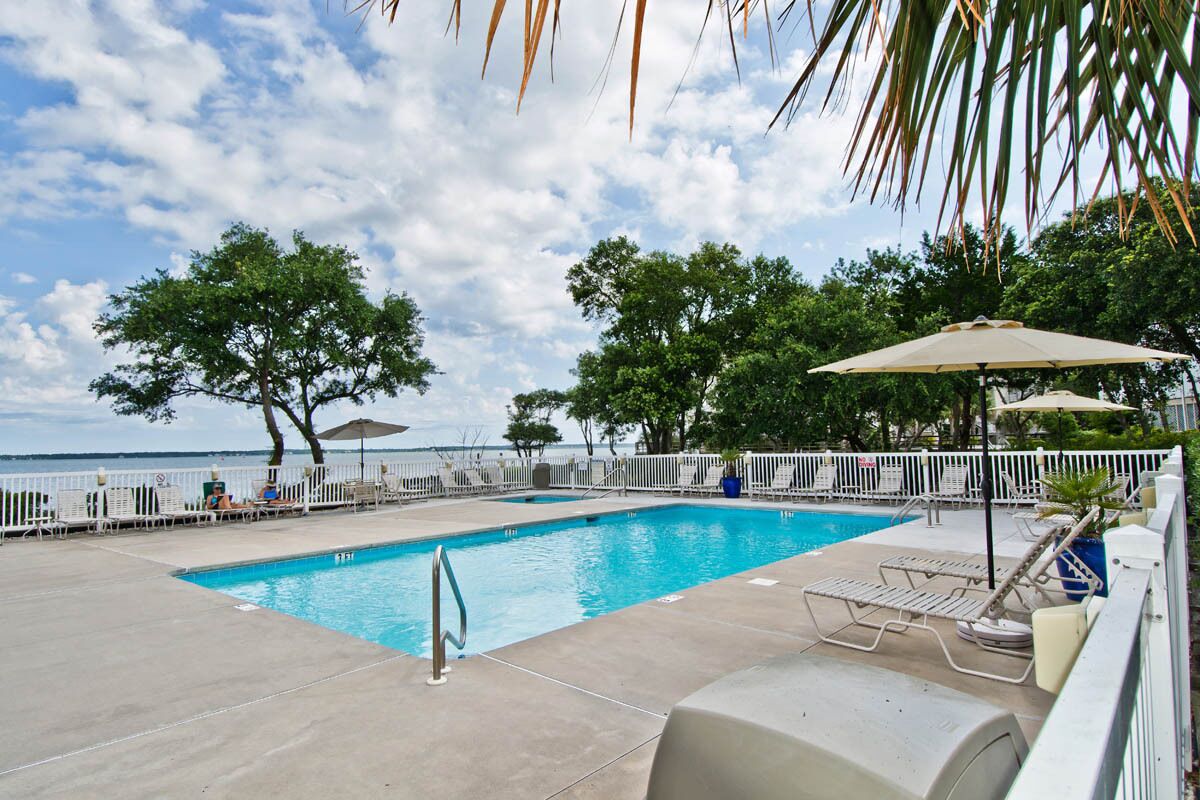 Une piscine rectangulaire avec un escalier en métal, entourée de chaises longues blanches et de parasols, avec des arbres et une vue sur la mer en arrière-plan.
