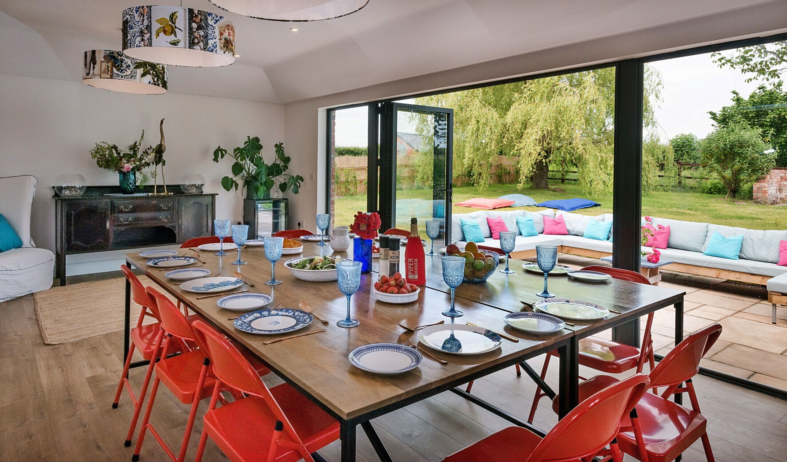A dining area with a large table set for six, surrounded by red chairs. There is a sofa with colorful cushions in the background, and a view of a garden through large windows.