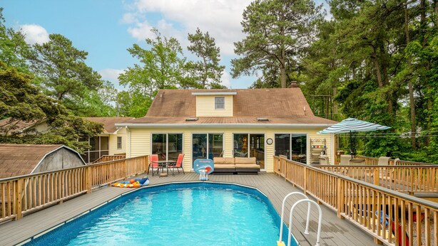 Ein Garten mit Schwimmbad, Holzterrasse, überdachter Terrasse und Gartenmöbeln, sowie ein Haus im Hintergrund.