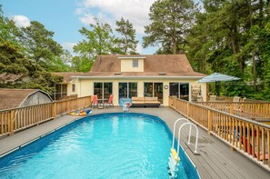 A backyard swimming pool with a wooden deck, a covered patio with outdoor furniture, and a house in the background. - Whelks on Willow by Seaside Vacations & Sales (Chincoteague Island)