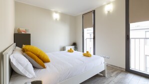 Een moderne slaapkamer met een groot bed, witte beddengoed en gele kussens. Er staat een nachtkastje met een lamp en een kleine plant. De kamer heeft een glazen schuifdeur die leidt naar een balkon.