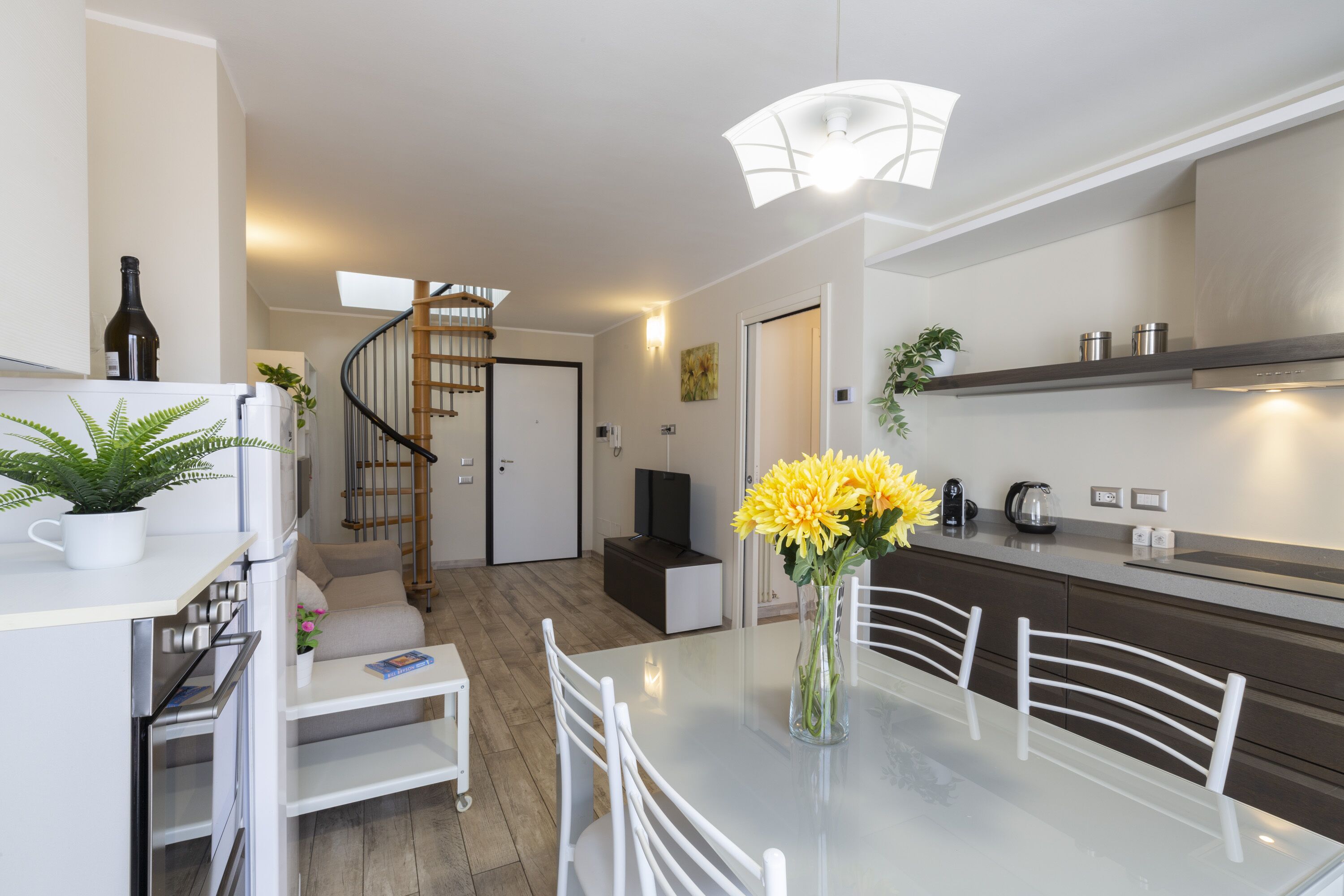 Una cocina moderna con comedor, que incluye una mesa de vidrio, sillas blancas y un jarrón con flores amarillas.