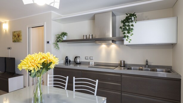 Een moderne keuken met een eethoek, met een witte tafel, twee witte stoelen, een vaas met gele bloemen, een roestvrijstalen spoelbak en een inbouw afzuigkap.