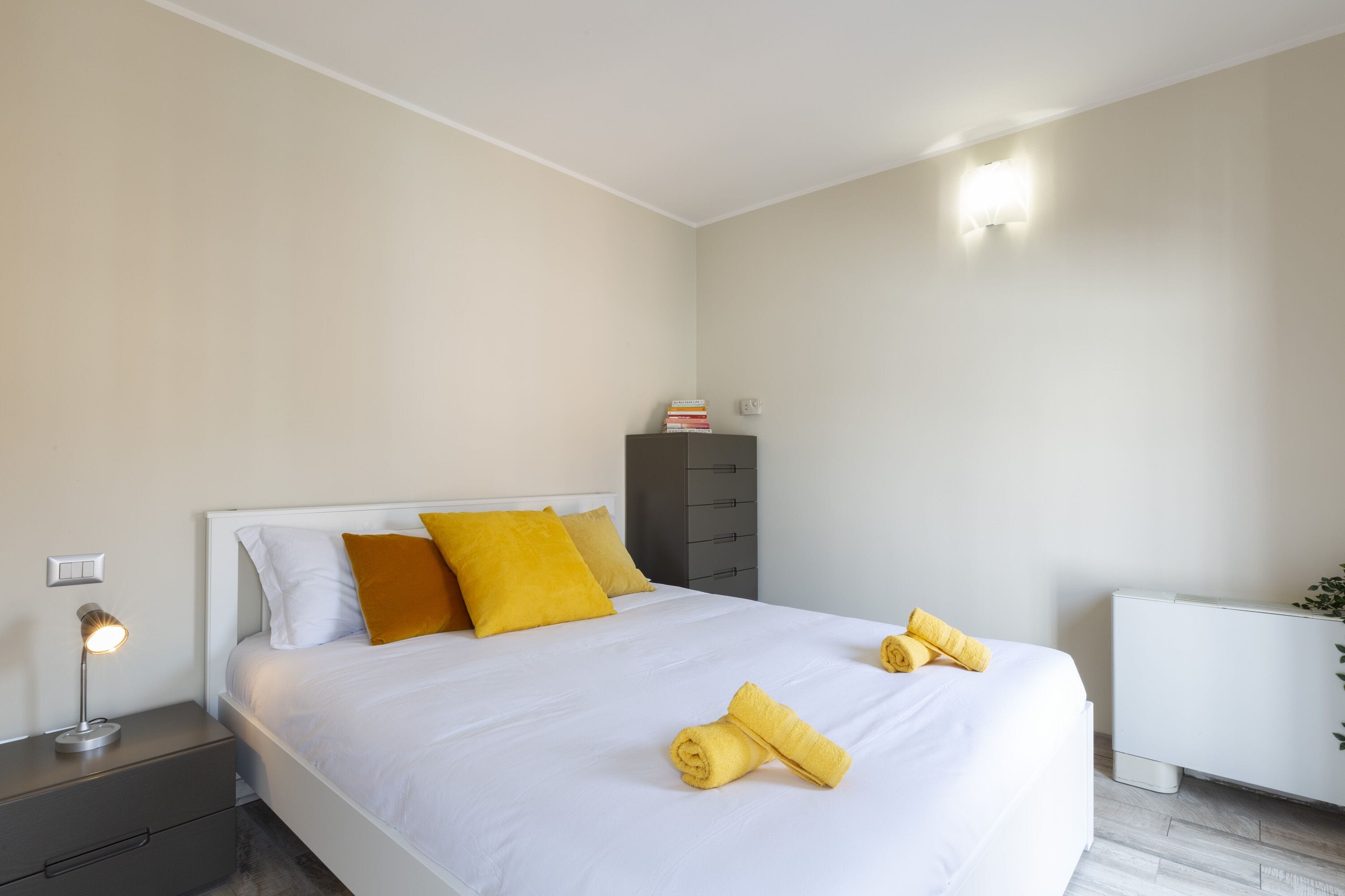 Un lit bien fait, avec une literie blanche et jaune, une lampe de chevet et une petite table de chevet dotée de tiroirs.