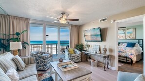 Ein geräumiges Wohnzimmer mit großem Fenster mit Blick auf den Strand, Deckenventilator, Flachbildfernseher und eine gemütliche Sitzecke mit einem Bett im Hintergrund.