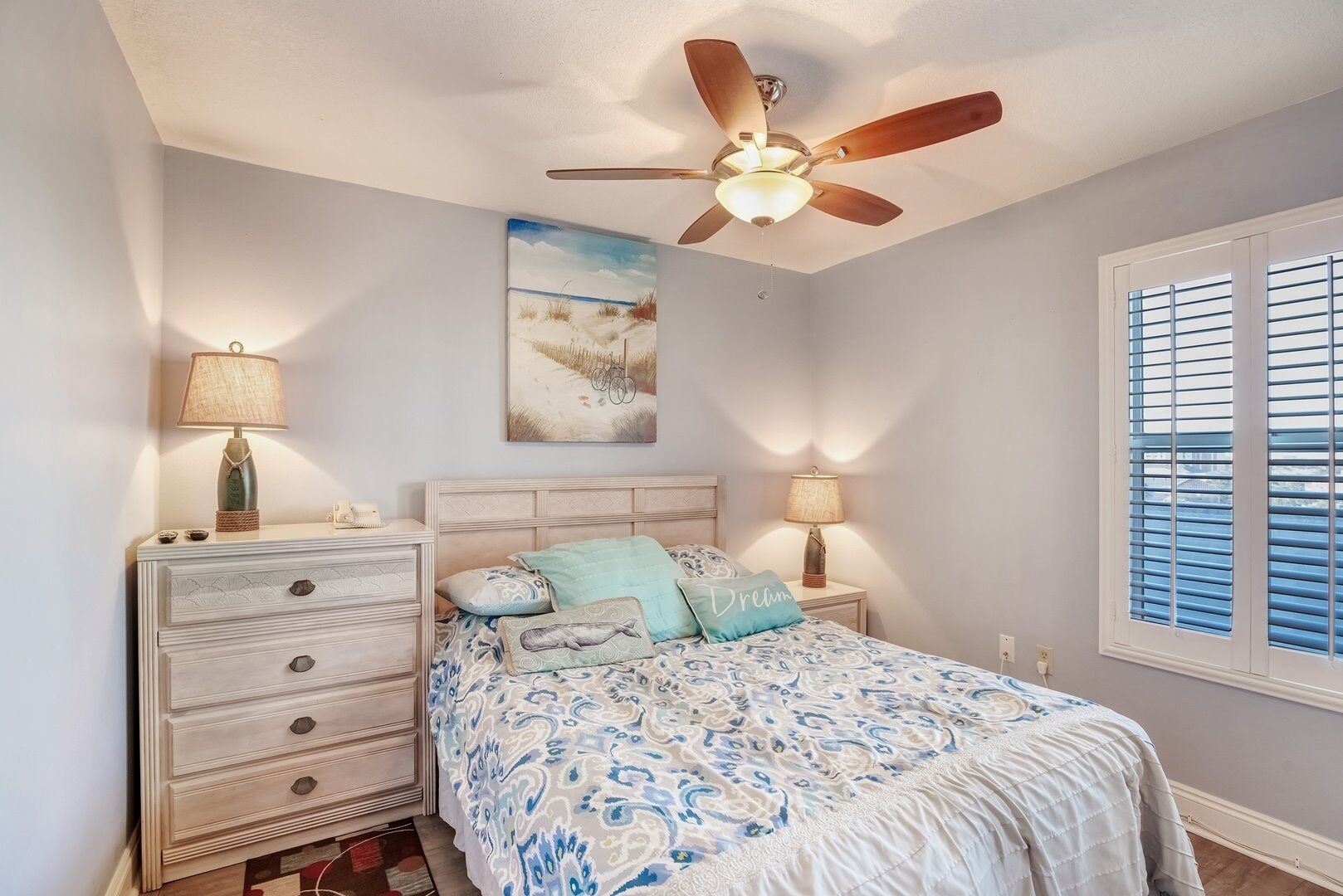 A bedroom with a bed, two bedside tables with lamps, a ceiling fan, a window with blinds, and a painting on the wall.