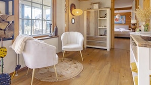 A cozy room with a large window, a white chair, a wooden floor, and a rustic bookshelf.