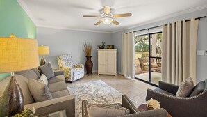 Ein Wohnzimmer mit Deckenventilator, einer Couch, einem Sessel, einem Schrank und Blick ins Freie.