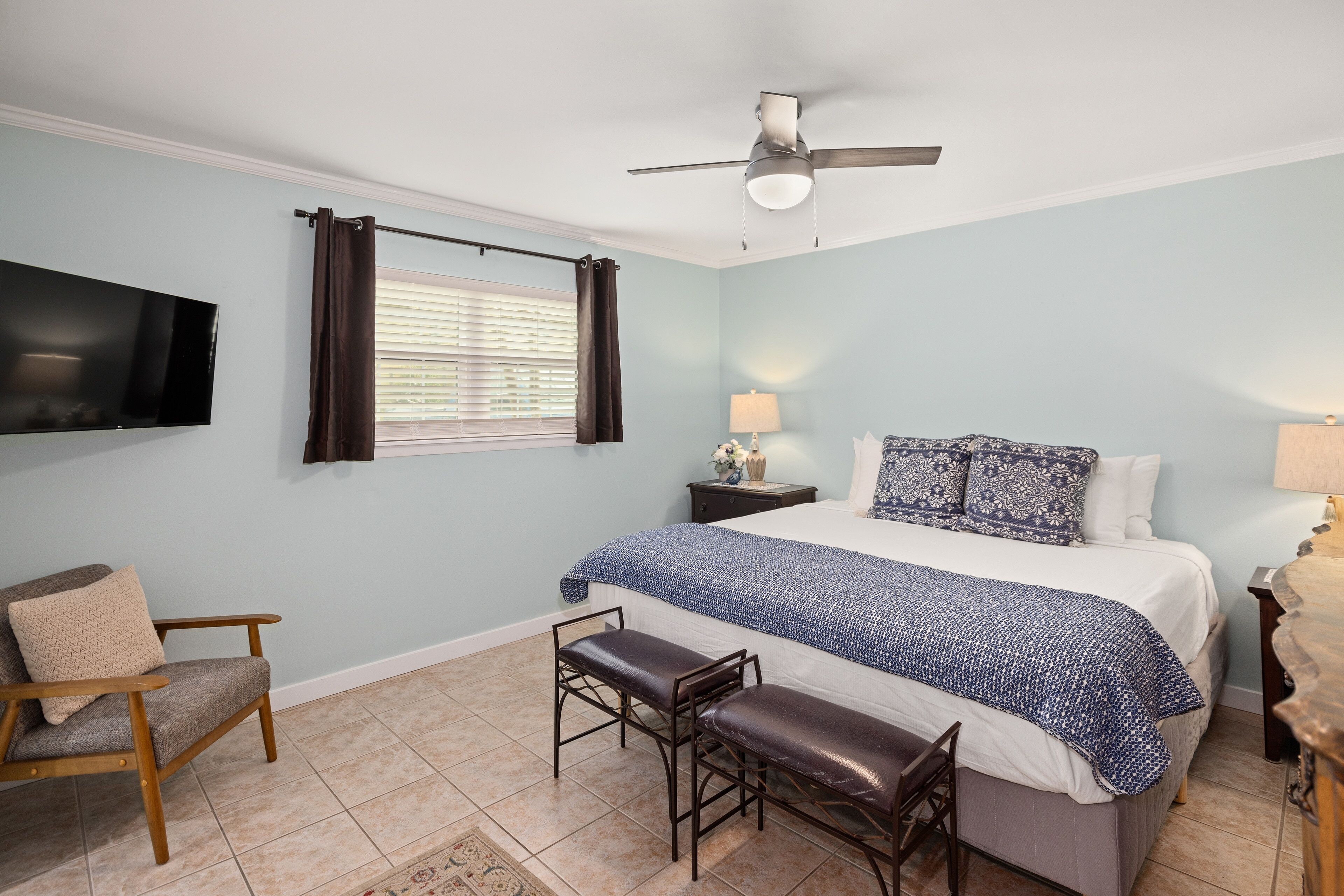 A bedroom with a bed, two chairs, a TV, a ceiling fan, and a window with curtains.